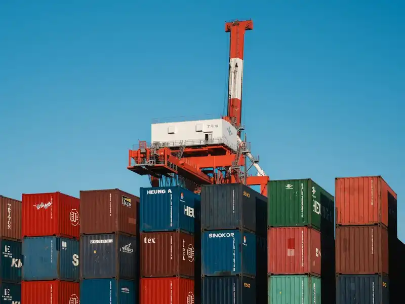 Shipping containers stacked several high at a port under a gantry crane, marked with Asian shipping lines including Heung-A, SINOKOR and KMTC